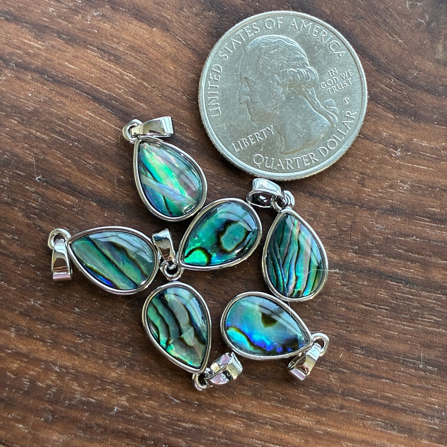 Four abalone shell Teardrop pendants with silver settings on a wooden surface next to a U.S. quarter dollar.