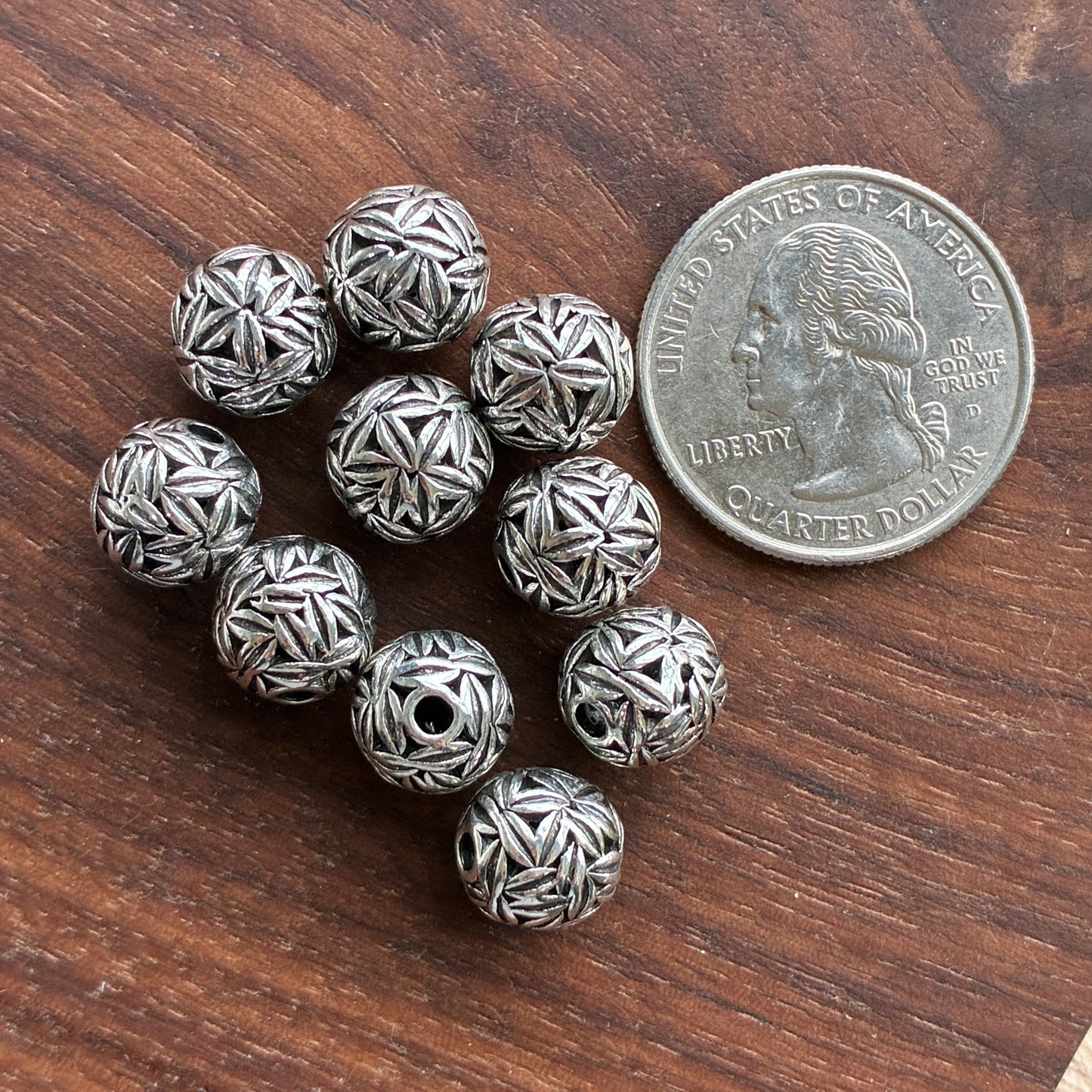 Silver beads with intricate patterns on a wooden surface next to a quarter for size comparison.