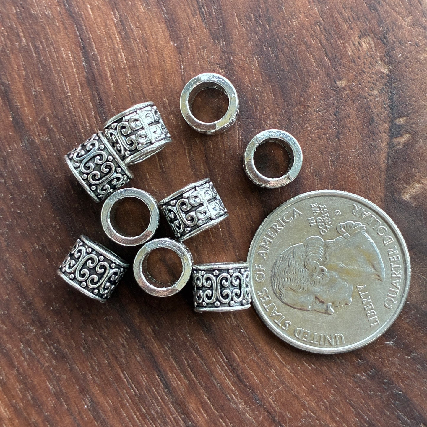 Silver beads with intricate patterns on a wooden surface next to a quarter for scale.