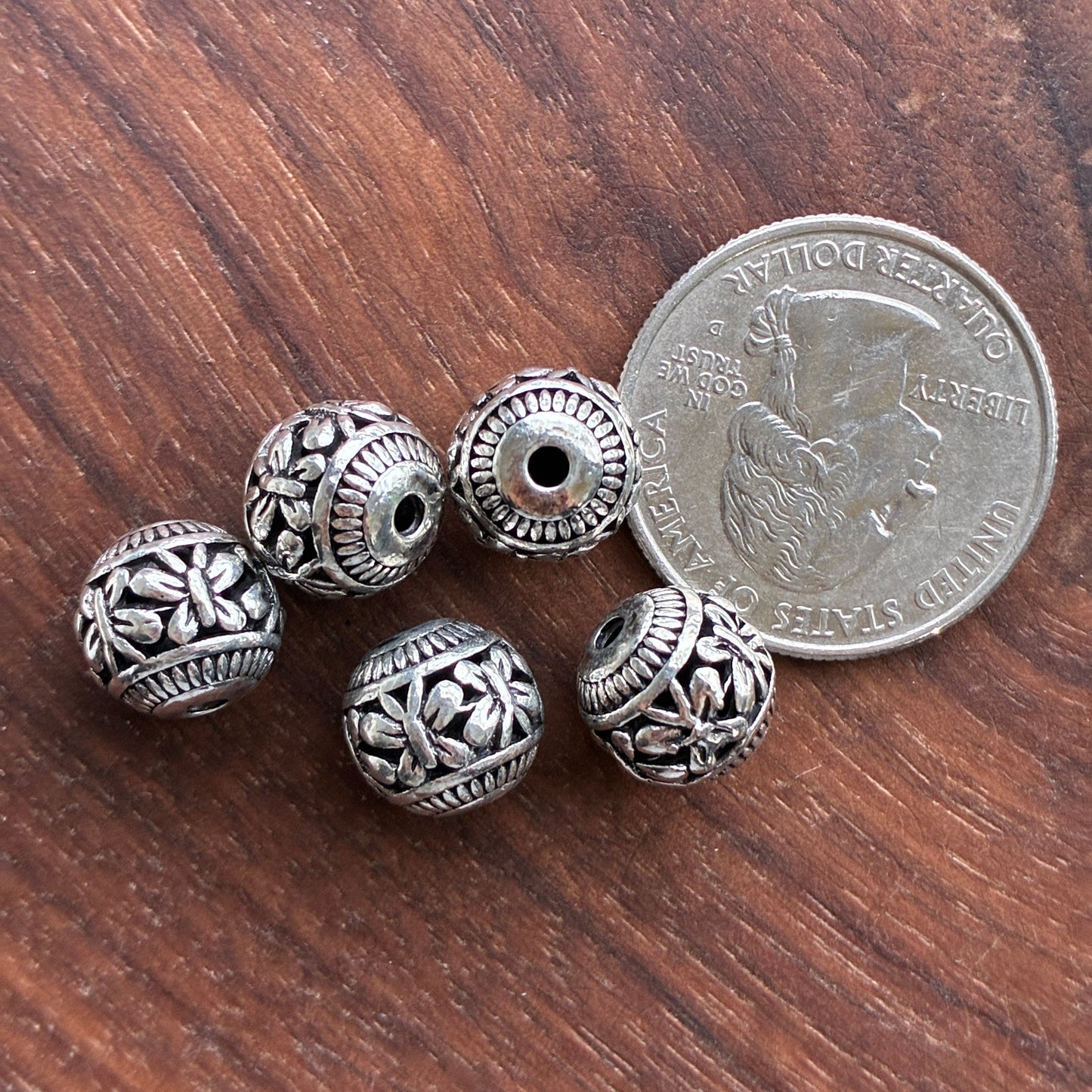 Silver beads with intricate designs on a wooden surface next to a U.S. quarter.