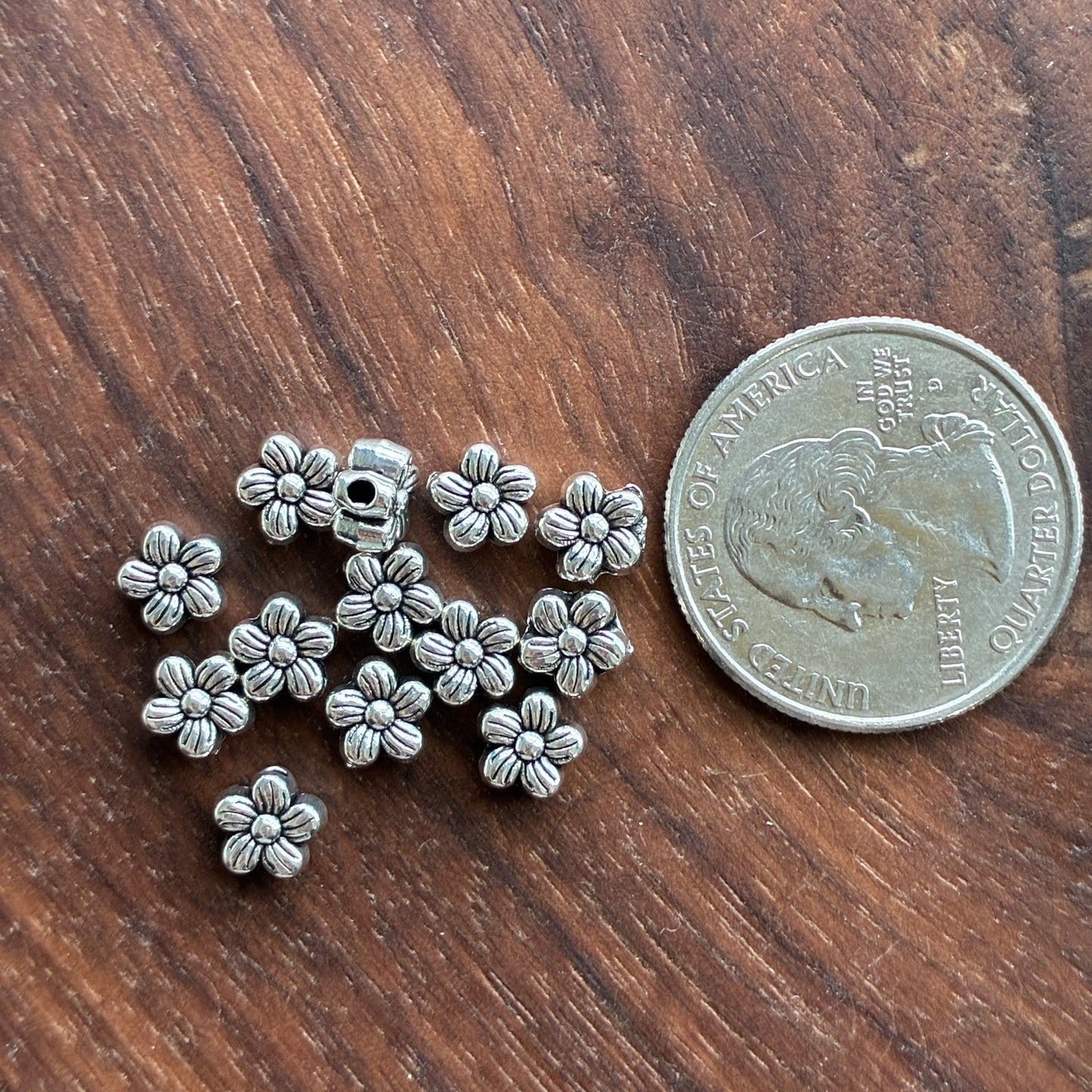Silver flower-shaped beads next to a U.S. quarter on a wooden surface